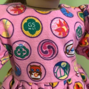 May include: Pink dress with a pattern of colourful circular designs. The dress features a round neckline and puffy sleeves. One of the circles has the text "GS WAY" in green. The dress is made of a soft, textured fabric.