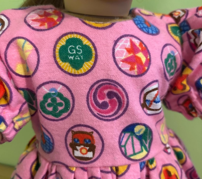 May include: Pink dress with a pattern of colourful circular designs. The dress features a round neckline and puffy sleeves. One of the circles has the text "GS WAY" in green. The dress is made of a soft, textured fabric.
