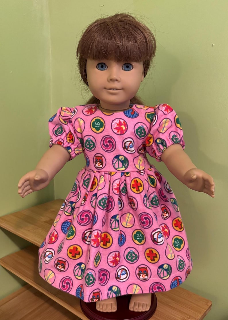May include: A doll in a pink dress patterned with colourful circular designs. The dress features short, puffed sleeves and a gathered skirt. The doll has brown hair and blue eyes.
