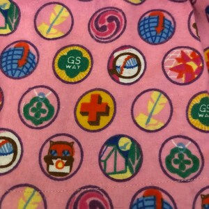 May include: Pink fabric patterned with circular badges. The badges display diverse designs, such as a cross, owl, feather, and globe. Some badges feature the text "GS WAY" in green and yellow.