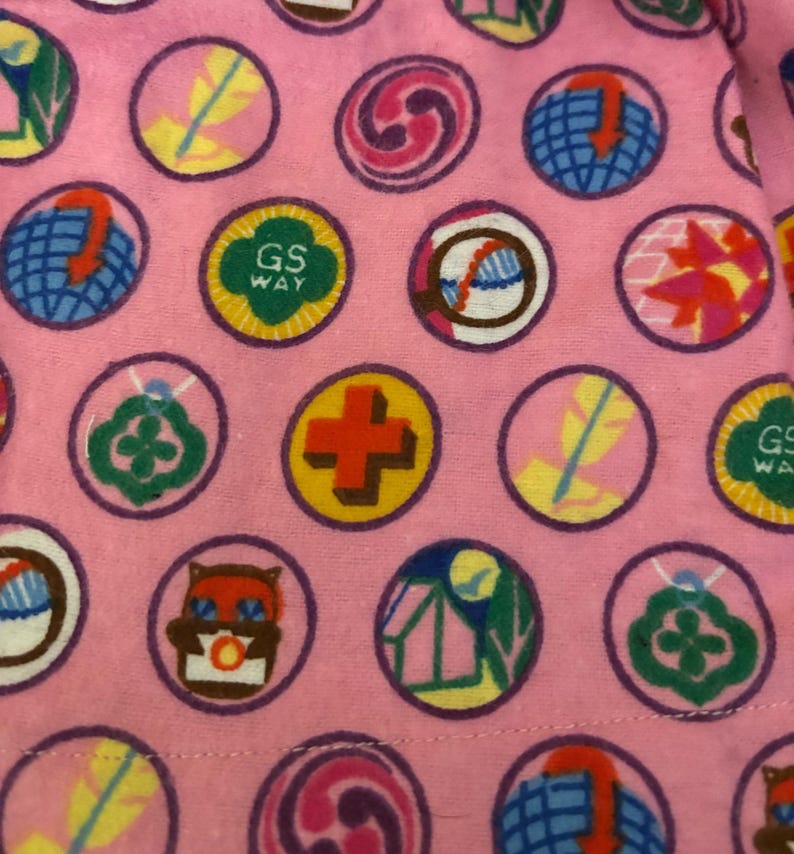 May include: Pink fabric patterned with circular badges. The badges display diverse designs, such as a cross, owl, feather, and globe. Some badges feature the text "GS WAY" in green and yellow.