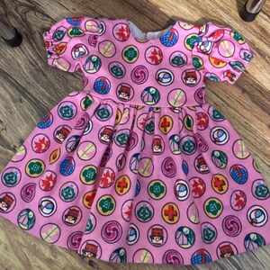May include: A pink dress with short, puffed sleeves and a full skirt. The dress features a repeating pattern of colourful circular designs, including the text "GS WAY". The dress is made of a soft fabric.