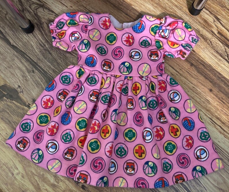 May include: A pink dress with short, puffed sleeves and a full skirt. The dress features a repeating pattern of colourful circular designs, including the text "GS WAY". The dress is made of a soft fabric.