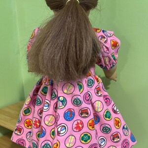 May include: A doll in a pink dress patterned with colourful circular designs. The dress features short sleeves and a flared skirt. The doll has brown hair tied back in a ponytail.