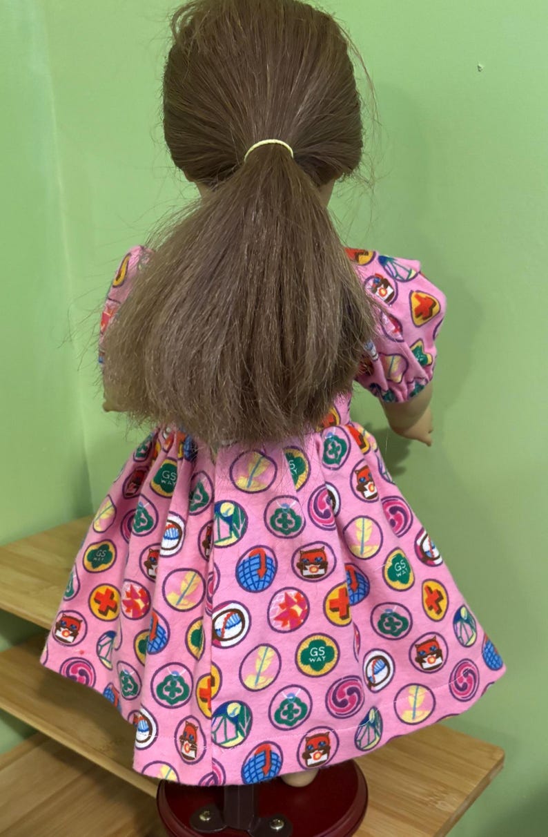 May include: A doll in a pink dress patterned with colourful circular designs. The dress features short sleeves and a flared skirt. The doll has brown hair tied back in a ponytail.