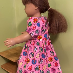 May include: A doll in a pink dress patterned with colourful circular designs. The dress features short sleeves and a gathered skirt. The doll has brown hair tied back in a ponytail.