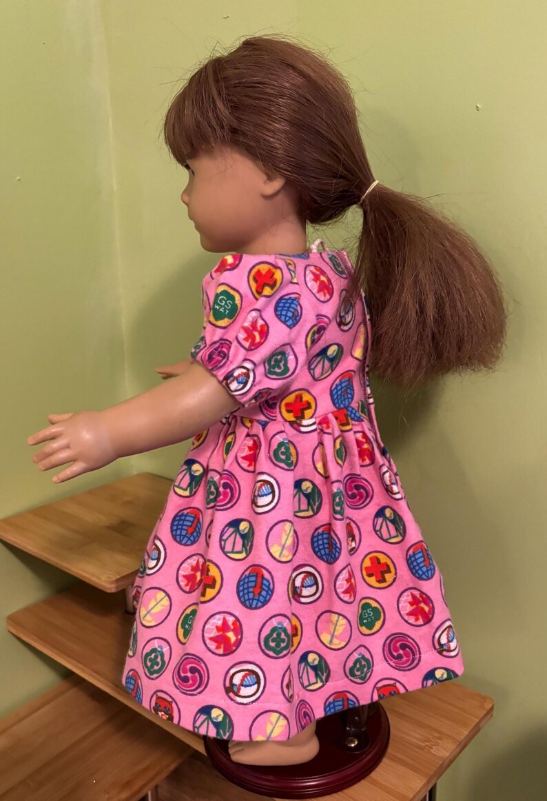 May include: A doll in a pink dress patterned with colourful circular designs. The dress features short sleeves and a gathered skirt. The doll has brown hair tied back in a ponytail.