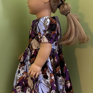 Może przedstawiać: Lalka z blond włosami zaplecionymi w warkocz na boku, ubrana w sukienkę z kr&oacute;tkim rękawem z powtarzającym się wzorem postaci animowanych, w tym bałwana, renifera oraz ludzi w fioletowych, niebieskich i brązowych strojach na ciemnym tle.