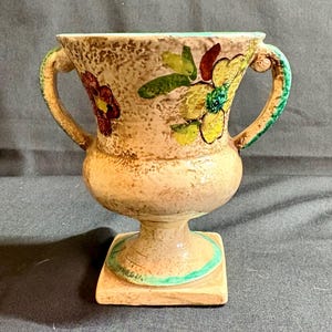 Könnte beinhalten: Eine Vintage-Keramikvase mit zwei Griffen, mit einer strukturierten beigefarbenen Oberfläche und floralen Designs in Gelb, Rot und Grün. Die Vase hat einen quadratischen Sockel mit grüner Umrandung. Ein dekoratives Stück für die Inneneinrichtung.