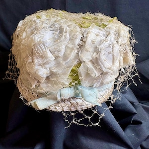 May include: Vintage white hat with a delicate net overlay. The hat features white fabric flowers and a light blue ribbon. The base of the hat is a woven material. A charming accessory for special occasions.