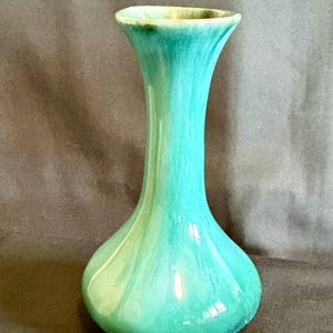 Puede incluir: Un jarrón de cerámica turquesa con base bulbosa y cuello largo y delgado. El jarrón tiene un acabado brillante y un borde acampanado. El color pasa de un verde azulado oscuro en la base a un turquesa más claro y brillante hacia la parte superior.