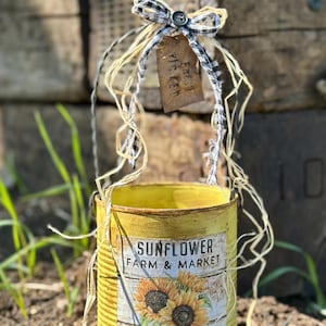 Może przedstawiać: Żółta puszka z motywem słonecznika, z napisem "Sunflower Farm & Market" i "Seeds & Flowers for Sale". Posiada czarno-białą kratkowaną kokardę i etykietę z napisem "Fresh Picked".