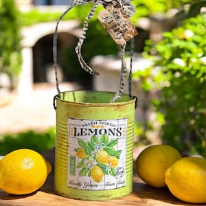 Könnte beinhalten: Ein grüner Metalleimer im Used-Look mit Henkel, verziert mit einer Schleife und einem Anhänger, zeigt "FRESH PICKED LEMONS" mit einer Zitronenillustration. Zwei leuchtend gelbe Zitronen liegen neben dem Eimer auf einer Holzoberfläche, was auf frische Produkte und Heimdekoration hindeutet.