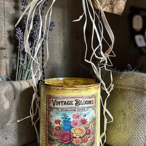 Puede incluir: Un recipiente de metal amarillo de estilo vintage con una etiqueta de paquete de semillas florales. La etiqueta dice "Vintage Blooms Heirloom Seeds" y "Mixed Varieties". El recipiente tiene un asa de alambre, rafia y una pequeña etiqueta que dice "Happy Spring".