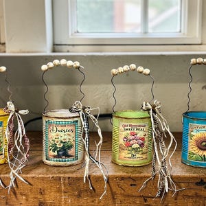 May include: Four decorative tin can lanterns with wire handles and beaded tops. Each can features a vintage seed packet design with floral illustrations. The cans are yellow, white, green, and blue, and are adorned with ribbons.