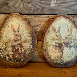 May include: Two oval wooden plaques with painted Easter bunny scenes. One shows a bunny with a basket of eggs, the other depicts two bunnies holding baskets. The plaques have a rustic, natural wood edge.