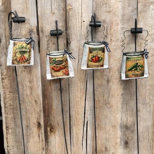 Può includere: Quattro paletti da giardino in metallo con pacchetti di semi in stile vintage attaccati. I pacchetti presentano illustrazioni di carote, pomodori, peperoni e cetrioli. Ogni pacchetto ha la scritta "SEEDS" stampata.