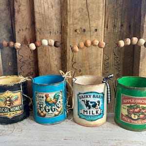 Może przedstawiać: Cztery metalowe puszki z różnymi wzorami i etykietami. Puszki są pomalowane w różnych kolorach, w tym czarny, niebieski, biały i zielony. Każda puszka ma druciany uchwyt z drewnianymi koralikami. Etykiety przedstawiają obrazy i tekst, w tym "Local Honey", "Farm Fresh Eggs", "Dairy Barn Milk" i "Apple Orchard Pick Your Own".