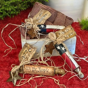May include: A patriotic-themed display featuring decorative wooden rolling pins painted in red, white, and blue, with star accents. The display includes tags with phrases like "God Bless America" and "We the People," all arranged in a rustic wooden box.