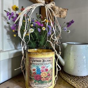 Może przedstawiać: Żółty metalowy pojemnik w stylu vintage wypełniony kolorowymi dzikimi kwiatami. Pojemnik ma napis "VINTAGE BLOOMS" i "HEIRLOOM SEEDS". Etykieta z napisem "Happy Spring" wisi na uchwycie. Przedmiot dekoracyjny.