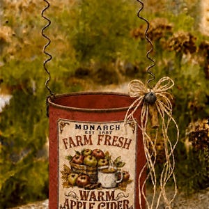 May include: A cylindrical red metal container with a wire handle featuring five wooden beads. The label reads MONARCH EST 1887 FARM FRESH WARM APPLE CIDER MADE WITH FRESH APPLES CINNAMON SPICE EVERYTHING NICE with an illustration of apples and a mug.