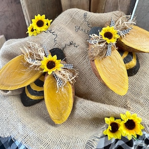 May include: Two wooden bee figurines with yellow and black paint, decorated with sunflowers and black and white ribbon. The bees are sitting on a burlap surface.