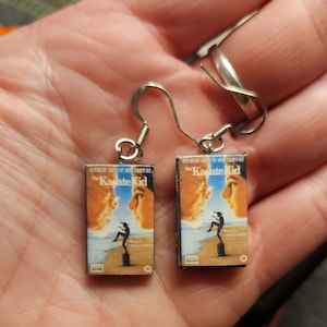 May include: A pair of silver earrings with a miniature The Karate Kid VHS tape charm on each earring. The charms feature the movie poster with the text "The Karate Kid" and a picture of Mr. Miyagi and Daniel LaRusso.