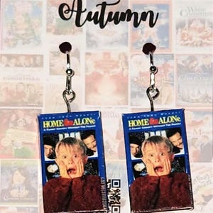 May include: A pair of earrings featuring miniature replicas of the Home Alone VHS movie cover. The earrings are silver and have a heart-shaped hook. The movie cover features a blue background with the title "Home Alone" in white text. The image shows a young boy with a shocked expression on his face.
