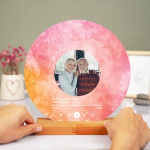 Könnte beinhalten: Runder, beleuchteter Foto-Display mit einem Paar in einem zentralen kreisförmigen Foto. Der Hintergrund hat einen Aquarell-Effekt in Rosa- und Orangetönen. Das Display enthält Songinformationen und einen Holzsockel.