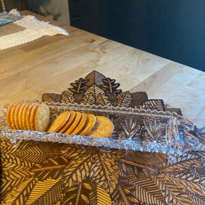 May include: A clear glass serving dish with a decorative pattern sits on a gold leaf patterned placemat. The dish is filled with a stack of round crackers.
