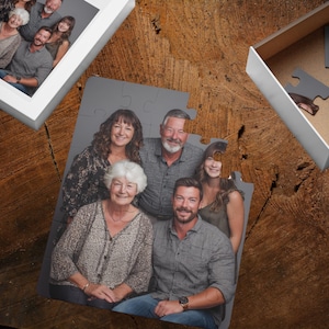 May include: A family portrait puzzle with a gray background, partially assembled on a wooden surface. The puzzle pieces show a group of six people smiling. A white box with puzzle pieces is in the background.