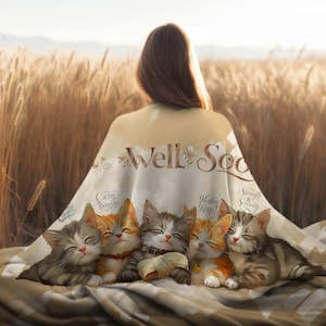 May include: A soft, beige blanket with a white background featuring a group of five cartoon cats with the text "Get Well Soon" in the center. The cats are all sleeping and have different expressions on their faces. The blanket is surrounded by a field of wheat.