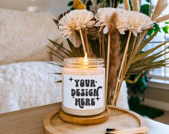 May include: A lit candle in a glass jar with a label that reads "YOUR DESIGN HERE." The candle sits on a wooden coaster and a round wooden tray. Dried flowers and matches are also visible.