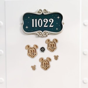 Puede incluir: Puerta blanca con un letrero decorativo que dice "11022" en letras blancas. Debajo del letrero hay varios recortes de madera con la cabeza de Mickey Mouse. El letrero tiene un fondo verde azulado con estrellas blancas.