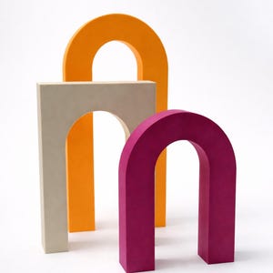 Op de afbeelding: Drie boogvormige decoratieve objecten in oranje, beige en magenta. De objecten staan rechtop tegen een witte achtergrond. De vormen zijn gemaakt van een massief materiaal. Studio-opname.