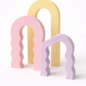 Op de afbeelding: Drie pastelkleurige boogsculpturen. De grootste is roze, de middelste is geel en de kleinste is lavendel. Elke boog heeft een golvend, gestructureerd ontwerp aan de zijkanten, wat een moderne, artistieke esthetiek creëert.