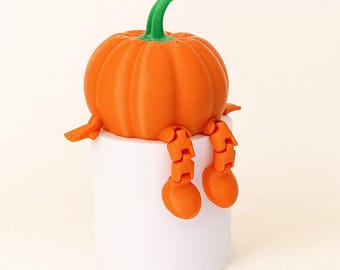 May include: An orange pumpkin figurine with a green stem and articulated orange legs, sitting on a white cylindrical base. The pumpkin has a ribbed texture, and the legs are posed as if dangling.