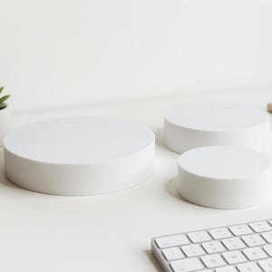 Pode incluir: Três plataformas de exibição circulares brancas de tamanhos variados são dispostas sobre uma superfície branca. Uma pequena planta suculenta em vaso está à esquerda. Um teclado e um caderno estão ao fundo.