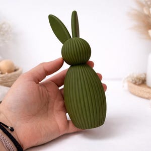 Pu&ograve; includere: Una statuetta decorativa di coniglietto pasquale verde con corpo a coste e lunghe orecchie. Il coniglietto &egrave; tenuto in mano, con sfondo bianco e altre decorazioni pasquali.