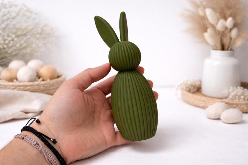 Pu&ograve; includere: Una statuetta decorativa di coniglietto pasquale verde con corpo a coste e lunghe orecchie. Il coniglietto &egrave; tenuto in mano, con sfondo bianco e altre decorazioni pasquali.