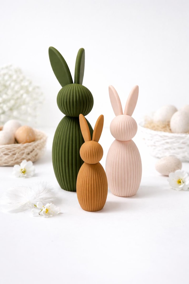 Pu&ograve; includere: Tre figurine decorative a forma di coniglio in verde oliva, pesca e rosa chiaro. I coniglietti hanno una consistenza a coste e orecchie lunghe. Fiori bianchi e uova in un cestino sullo sfondo, che suggeriscono un tema pasquale.