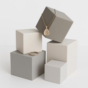 May include: A gold pendant necklace with a teardrop-shaped charm hangs from a gold chain. A gold ring with a textured band sits on a gray cube. The jewelry is displayed on a collection of gray and white cubes.