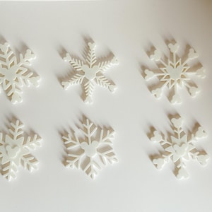 May include: Six white snowflake ornaments with a Mickey Mouse head design in the center. The ornaments are arranged on a white surface, casting soft shadows. These decorative pieces are ideal for winter holiday decor.