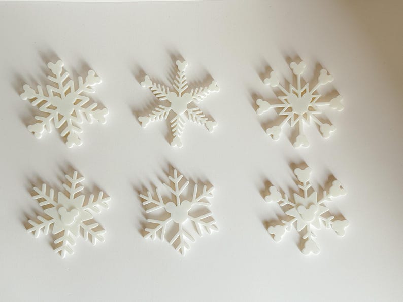May include: Six white snowflake ornaments with a Mickey Mouse head design in the center. The ornaments are arranged on a white surface, casting soft shadows. These decorative pieces are ideal for winter holiday decor.