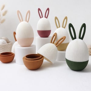 May include: Easter-themed decorative egg-shaped containers with bunny ear accents. The eggs are white with ribbed detailing and come in various colors, including pink, burgundy, yellow, and green. Some are open, revealing a matching interior.