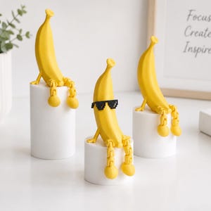 Könnte beinhalten: Drei gelbe Bananenfiguren mit beweglichen Gliedmaßen, die jeweils auf einem weißen zylindrischen Sockel sitzen. Die mittlere Bananenfigur trägt eine schwarze, verpixelte Sonnenbrille, während die anderen beiden ein schlichtes gelbes Gesicht haben.