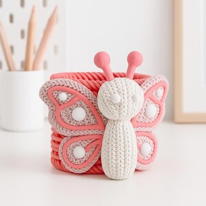 May include: A decorative pink and white butterfly-shaped basket. The basket has a woven texture with a butterfly design on the front. The butterfly has a white knitted body and pink wings with white accents. The basket is likely used for storage or decoration.