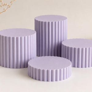 Può includere: Un set di quattro piedistalli espositivi cilindrici di colore lavanda chiaro. Ogni piedistallo presenta una texture verticale scanalata o a coste lungo la circonferenza esterna, con piani circolari piatti di altezze variabili per l'esposizione.