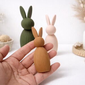 Pu&ograve; includere: Una mano tiene in mano una piccola statuetta di coniglio marrone a coste. Dietro ci sono due statuette di coniglio pi&ugrave; grandi in verde oliva e rosa pallido. Le statuette sono decorative e adatte per la decorazione pasquale o primaverile.