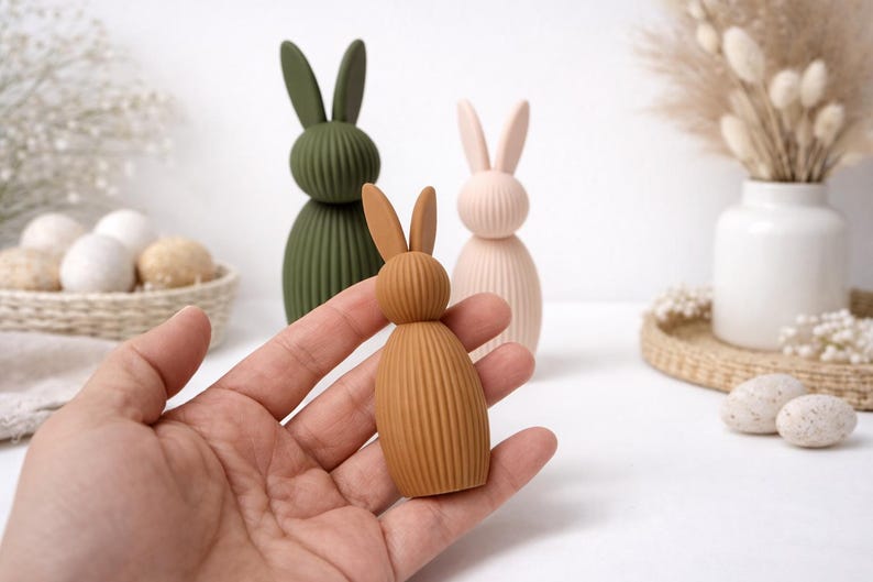 Pu&ograve; includere: Una mano tiene in mano una piccola statuetta di coniglio marrone a coste. Dietro ci sono due statuette di coniglio pi&ugrave; grandi in verde oliva e rosa pallido. Le statuette sono decorative e adatte per la decorazione pasquale o primaverile.